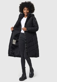 Manteau long noir matelassé avec capuche et fermeture éclair, doté d'un design matelassé, de poches latérales et d'un tissu texturé. Porté ouvert sur un haut blanc.