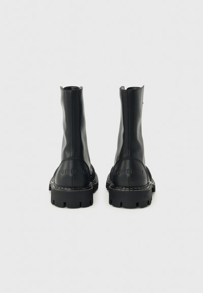 Ζευγάρι μαύρες δερμάτινες μπότες μέσης μοσχάρι με χοντρές σόλες λουγκ, εμφανή ραφές και ανάγλυφο όνομα μάρκας στη φτέρνα, που φαίνεται από πίσω.