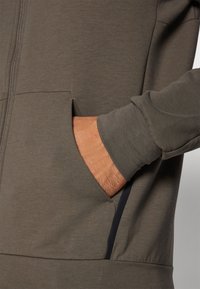 Sudadera con cremallera en verde oliva con un acabado texturizado. Presenta bolsillos laterales y acentos oscuros contrastantes a lo largo de las costuras. Incluye mangas largas.