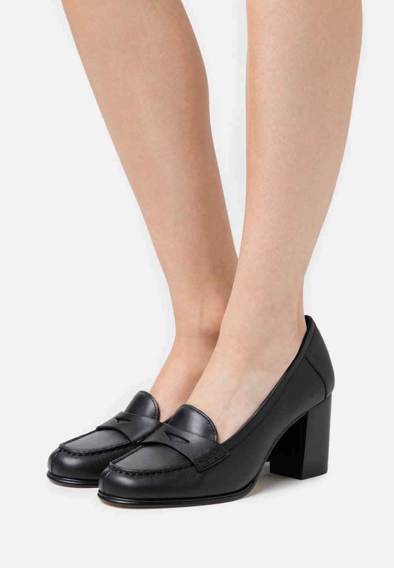 Mocassins à talons carrés en cuir noir portés sur des pieds nus, avec un bout arrondi et un détail de bride sur le dessus.
