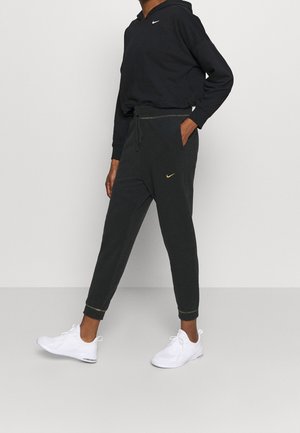 Person trägt schwarzen Nike-Hoodie, schwarze Nike-Jogginghose mit goldenem Logo und weiße Sportschuhe, steht mit einer Hand in der Tasche.
