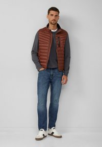Chaleco acolchado marrón sobre una camiseta de manga larga gris, combinado con jeans azules y zapatillas blancas. Bolsillo con cremallera en el chaleco.
