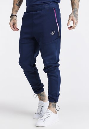 Mand iført marineblå joggingbukser med elastisk talje, lynlåslommer i siden og hvide sneakers, med tatoverede arme og ankler.