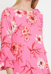 Nainen, jolla on päällään vaaleanpunainen mekko, jossa on punaisia ja valkoisia kukkakuoseja sekä rypytetyt kolmen neljänneksen hiukset, esittelee ylävartaloaan ja kaula-aukkoaan.