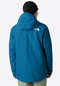 Un personaggio che indossa una giacca con cappuccio blu con il logo The North Face sul retro, abbinata a pantaloni chiari e un berretto nero.