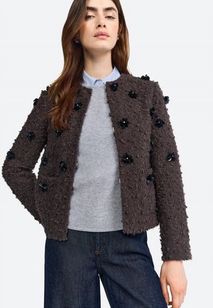 Chaqueta marrón texturizada con adornos florales, combinada con un suéter gris claro y jeans de mezclilla oscuros. Presenta un diseño cuadrado y bolsillos frontales.