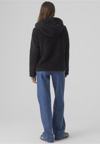 Person klädd i en svart huvtröja av fleece och blå jeans, står vänd bortåt mot en enfärgad ljusgrå bakgrund.