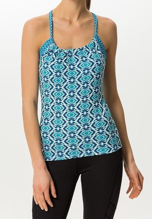 Kobieta ubrana w bezrękawnik w turkusowo-niebieskie geometryczne wzory oraz dopasowane czarne spodnie na jednolitym jasnym tle.