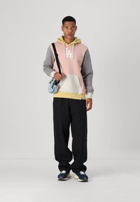 Sudadera con capucha en rosa, gris y amarillo con un logo blanco, combinada con pantalones negros holgados y zapatillas azul marino. Incluye una bolsa azul.