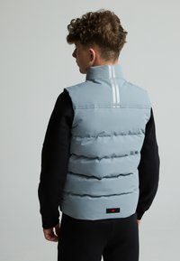 Gilet matelassé bleu clair avec col haut, présentant un design matelassé horizontal et deux bandes blanches verticales à l'arrière. Étiquette de marque noire à l'ourlet.