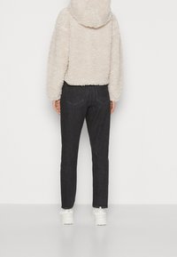 Personne portant une veste polaire crème, un jean droit noir et des baskets blanches épaisses, debout devant un fond blanc uni.