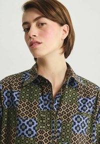 Chemise à boutons multicolore présentant un mélange de motifs floraux et géométriques dans des tons de bleu, vert et marron, avec un col et une poche sur la poitrine.