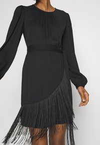 Femme portant une robe noire à manches longues avec une taille ajustée et des détails en franges le long de l'ourlet asymétrique.