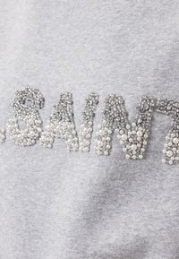 Grijze sweatshirt met getextureerde parelmoer letters in zilverachtige tonen en witte parels, die een verhoogd, gedetailleerd ontwerp creëren.