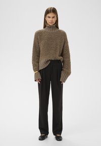 Smeđi teksturirani pulover s visokim ovratnikom, uparen s crnim širokim hlačama. Ima dugačke rukave s rebrastim manžetama i opušteni kroj.