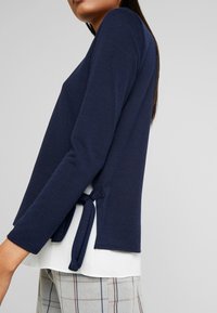 Marinblå stickad topp med långa ärmar och en knuten detalj på baksidan, lagd över en vit underdel, ihop med rutiga grå byxor.