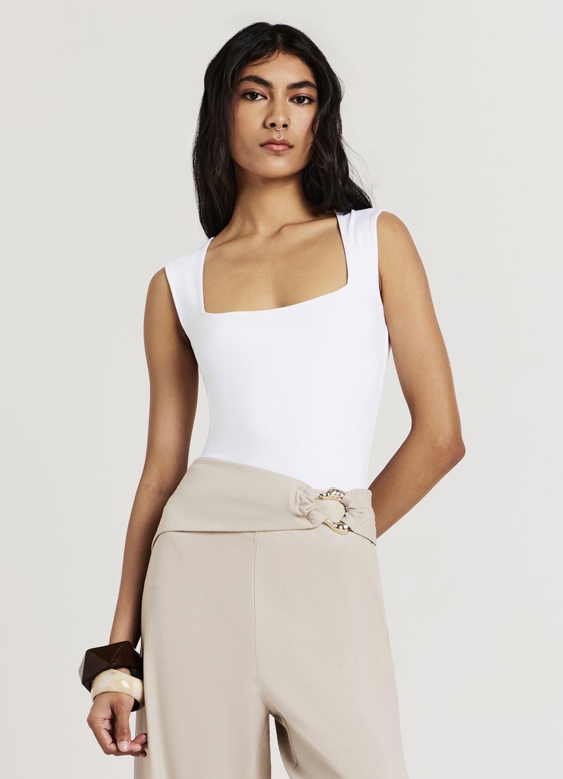 Body bianco con scollo quadrato abbinato a pantaloni beige a vita alta con cintura annodata e dettagli in metallo dorato. Bracciale marrone e crema al polso.
