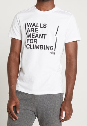 Mężczyzna w białej koszulce z czarnym napisem "Ściany są do wspinaczki" i szarych spodniach, stojący na tle jednolitego tła.