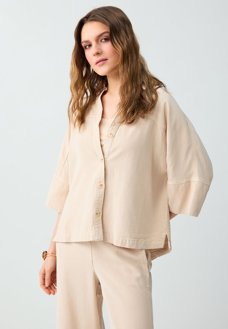 Femme aux cheveux ondulés portant une chemise beige ample boutonnée et un pantalon assorti, debout devant un fond clair uni.