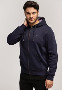 Granatowa bluza zapinana na zamek, wykonana z miękkiego materiału, z kieszenią z przodu, kapturem ze sznurkiem i haftowanym logo na lewej piersi.