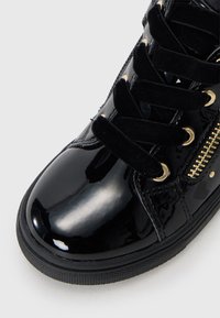 Fekete lakkbőr sneaker bársony fűzővel, arany fémlyukakkal és díszes oldalsó cipzárral. Texturált fekete gumitalppal rendelkezik.