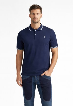 Uomo con polo blu navy con bordi bianchi e jeans blu a toppe, in piedi davanti a uno sfondo bianco semplice.
