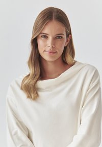 Fehér, túlságosan nagy, hosszú ujjú felső, bársonyos anyagból, csónaknyakkal. Sima textúra, minimalista dizájn, ejtett vállakkal.