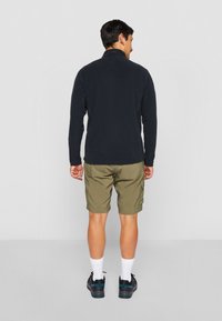 Veste en polaire noire avec un col montant, assortie à un short cargo vert olive. Portée avec des chaussettes blanches et des chaussures foncées avec des accents en teal.