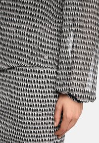 Grau meliertes Gewebe mit einem gepunkteten Muster in Schwarz und Weiß. Hat eine lockere Passform, geraffte Ärmel und ein geprägtes Metall-Detail.