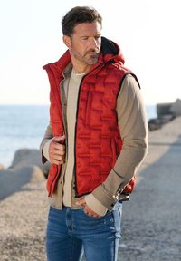 Rote gesteppte Steppweste, getragen über einem beigen Pullover. Mit Kapuze, Frontreißverschluss und Seitentaschen. Jeans runden das Outfit ab.