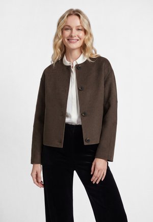 Smiling woman with blonde hair wearing a brown buttoned jacket over a white shirt and black pants against a plain background.