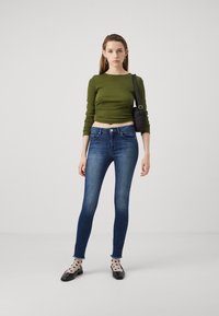 Grønn ribbete langermet topp, blå slim-fit jeans, prikkete sokker, svarte flate sko, liten svart veske. Jevne teksturer og tilpasset design.