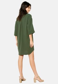 Loose-fitting, short-sleeve dress in olive green fabric, featuring a rounded hem and side slits, paired with beige heeled sandals.
