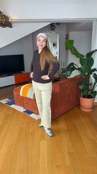 Νεαρή γυναίκα με μακριά μαλλιά, κορδέλα, σκούρο τοπ, ανοιχτόχρωμο παντελόνι με σχέδια αστεριών, στέκεται σε ένα καθιστικό κοντά σε ένα πορτοκαλί καναπέ και μεγάλο γλαστρικό φυτό.