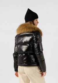 Schwarze Winterjacke aus Steppmaterial mit glänzender, genähter Textur und braunem Fellkragen, getragen mit einer schwarzen Mütze, von hinten betrachtet.