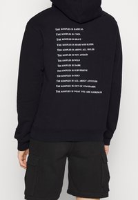 Sudadera negra con texto blanco en la parte posterior, que presenta frases repetidas sobre "The Kooples". Fabricada en un tejido suave, con un bolsillo tipo canguro y capucha.