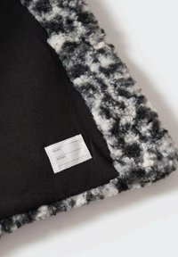 Chlupatá černobílá textilie s hladkým černým vnitřním podšívkou. Obsahuje malou bílou štítku na jméno a telefon.