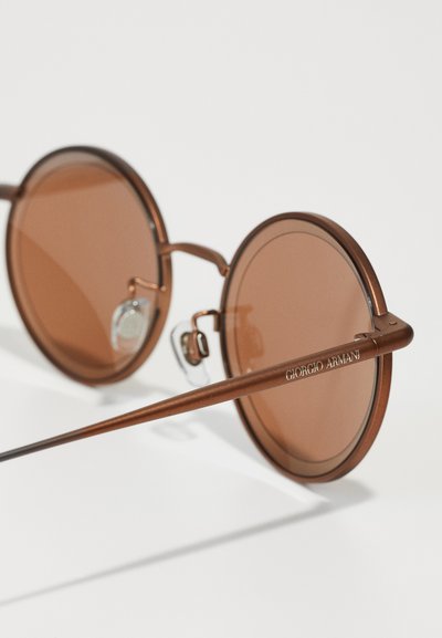 Round sunglasses with brown tinted lenses, metal frame, and sleek arms. Features clear nose pads and engraved brand name on the arm.