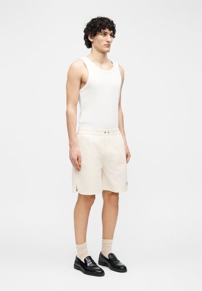 Jeune homme aux cheveux bouclés portant un débardeur blanc, un short beige, des chaussettes beiges et des mocassins noirs, debout devant un fond uni.