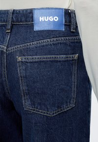 Vue arrière d'un jean en denim bleu foncé avec des surpiqûres jaunes et un patch en cuir bleu portant la mention "HUGO" au-dessus de la poche arrière.