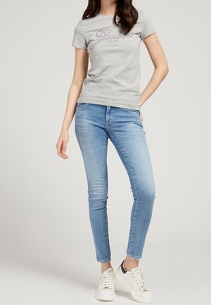 Kvinde iført grå Guess T-shirt, lyseblå skinny jeans og hvide sneakers, stående med den ene hånd på hoften mod en ensfarvet baggrund.