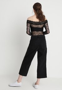 Musta pitsinen olkaimeton toppi yhdistettynä korkeavyötäröisiin mustiin leveälahkeisiin farkkuihin. Mallilla on valkoiset lenkkarit; näkyvissä pitsikuvio ja kontrastivärinen ompelu.