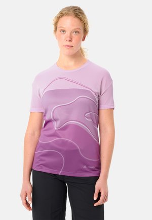 Junge Frau mit lockigem blondem Haar, die ein kurzärmeliges Shirt in lila Farbverlauf mit abstrakten Linienmustern und schwarze Hosen trägt, steht.