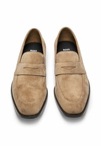 Beige suède loafers met een zachte textuur, voorzien van een decoratieve band en uitsnijding, en een donkerbruine rubberen zool.