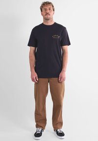 T-shirt bleu marine à col rond avec un logo cuivre sur la poitrine, associé à un chino beige et des baskets noires avec des accents blancs.