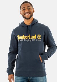 Navyblauwe katoenen hoodie met een voorkantzak, ribgebreide manchetten en een buidelzak. Het heeft gele Timberland-logo tekst en een boomgrafiek.