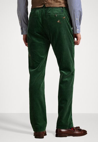 Green corduroy trousers with a vertical ribbed texture, back pockets, and embroidered accents. Paired with brown loafers, shirt sleeves visible.