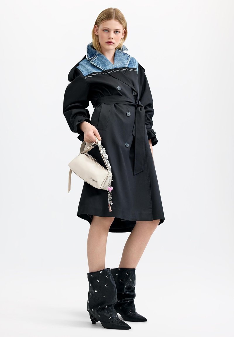 Woman wearing a black coat with a denim collar, holding a light beige handbag, and wearing black ankle boots with eyelet details.