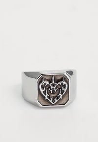 Silberner Metallring mit quadratischer Fläche, der ein erhabenes, kunstvolles Herzdesign vor einem strukturierten dunklen Hintergrund zeigt.
