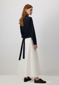 Blusa avvolgente blu navy con maniche lunghe, dettaglio con fiocco sul retro; abbinata a una gonna bianca fluida e scarpe marroni con lacci. Finitura in tessuto liscia.